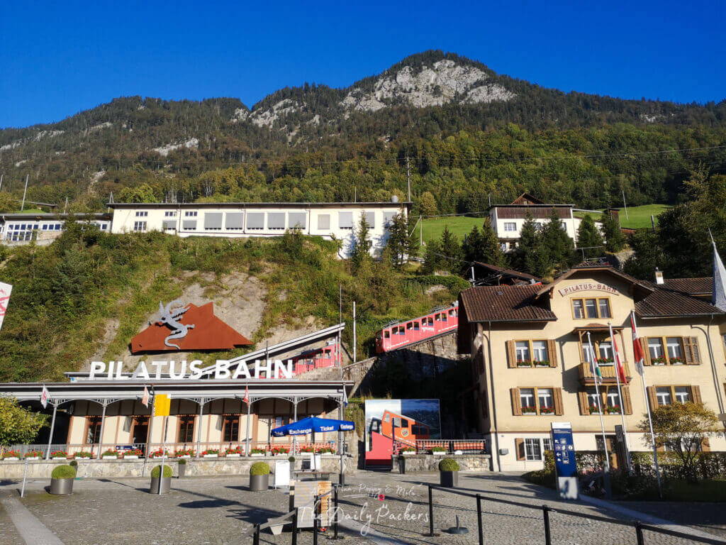 Entrée de la gare à crémaillère du Pilatus Bahn à Alpnachstad, avec le train rouge et la montagne verte en toile de fond.