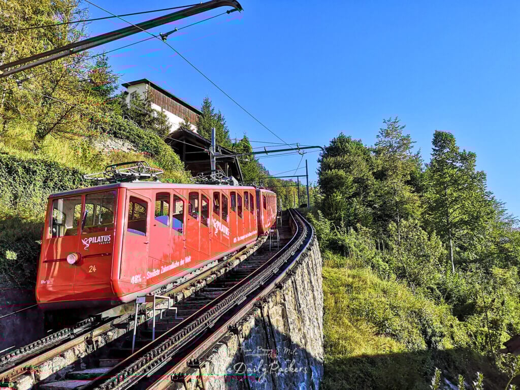 Train à crémaillère rouge gravissant les pistes escarpées du mont Pilatus, entouré de forêts et d'un ciel bleu limpide.