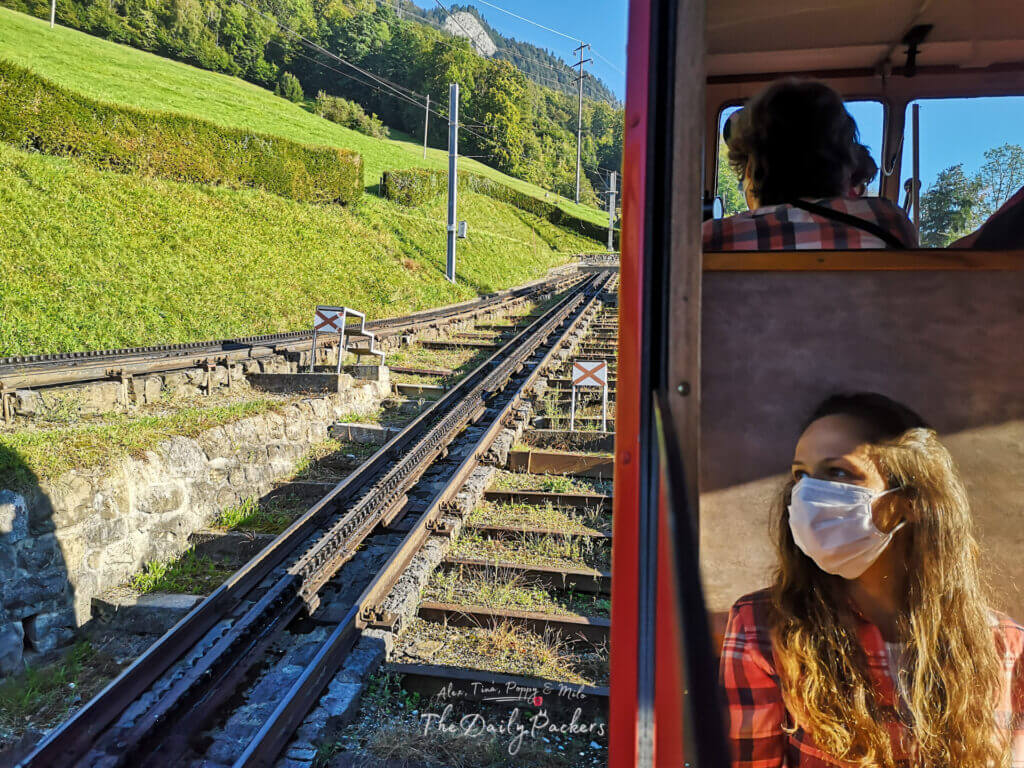 Femme portant un masque, assise à l'intérieur du train à crémaillère Pilatus, les rails étant visibles par la fenêtre.