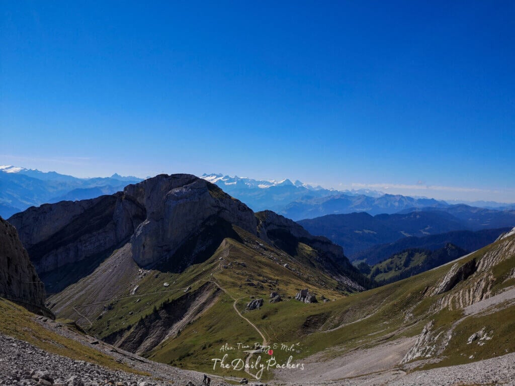 Randonneurs empruntant un étroit sentier de montagne menant au sommet du mont Pilatus, avec des vallées et des sommets au loin.