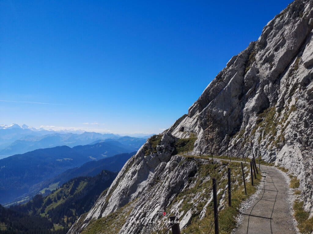 Sentier de randonnée étroit le long des pentes rocheuses du mont Pilatus, avec vue panoramique sur les vallées boisées et les Alpes au loin.