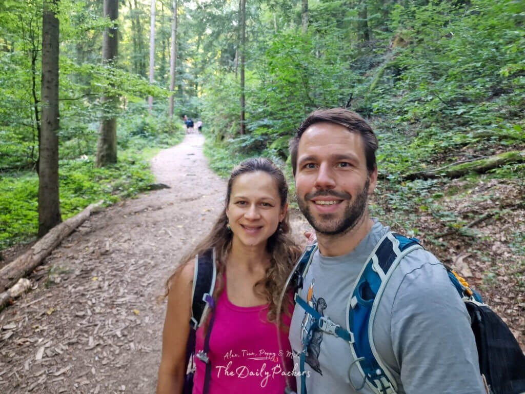 Couple souriant sur un sentier forestier à Sasbachwalden, entouré de grands arbres et de la douce lumière du soleil.