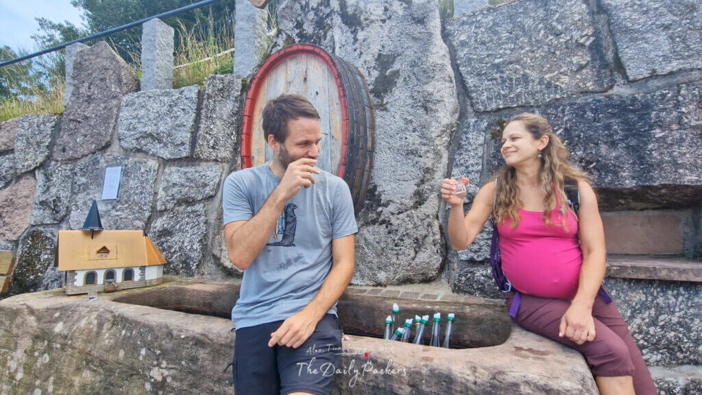Un couple partage un sourire en trinquant avec des schnaps à une fontaine en pierre construite dans un mur rocheux.