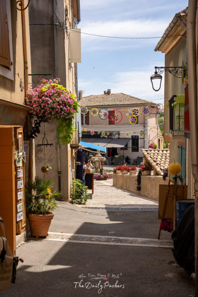 Ruelle étroite de Valensole menant à la place de la ville, bordée de fleurs et de vitrines colorées.