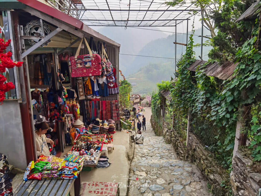 Narrow stone path lined with small market stalls in Cat Cat Village, selling colorful handicrafts, hats, and snacks with a view of the mountains ahead.
