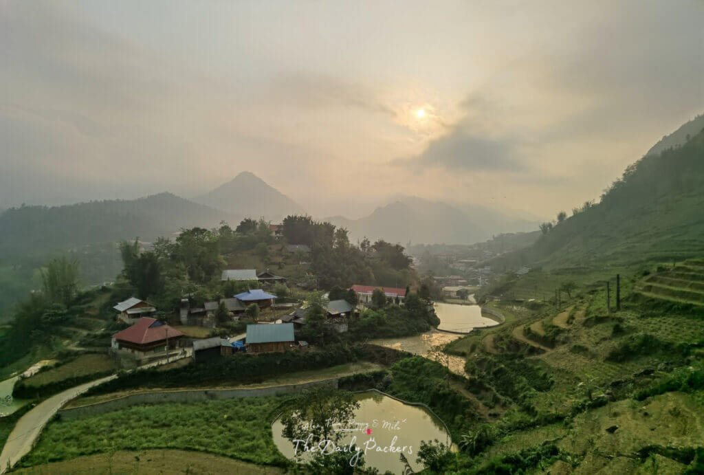 Scenic view of Cat Cat Village at sunset with rice terraces, small houses, winding paths, and misty mountains in the background.