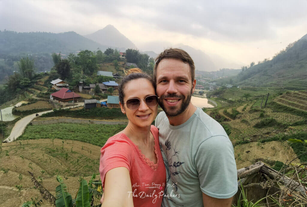 Couple taking a selfie with the green rice terraces, village houses, and misty mountains of Cat Cat Village behind them.