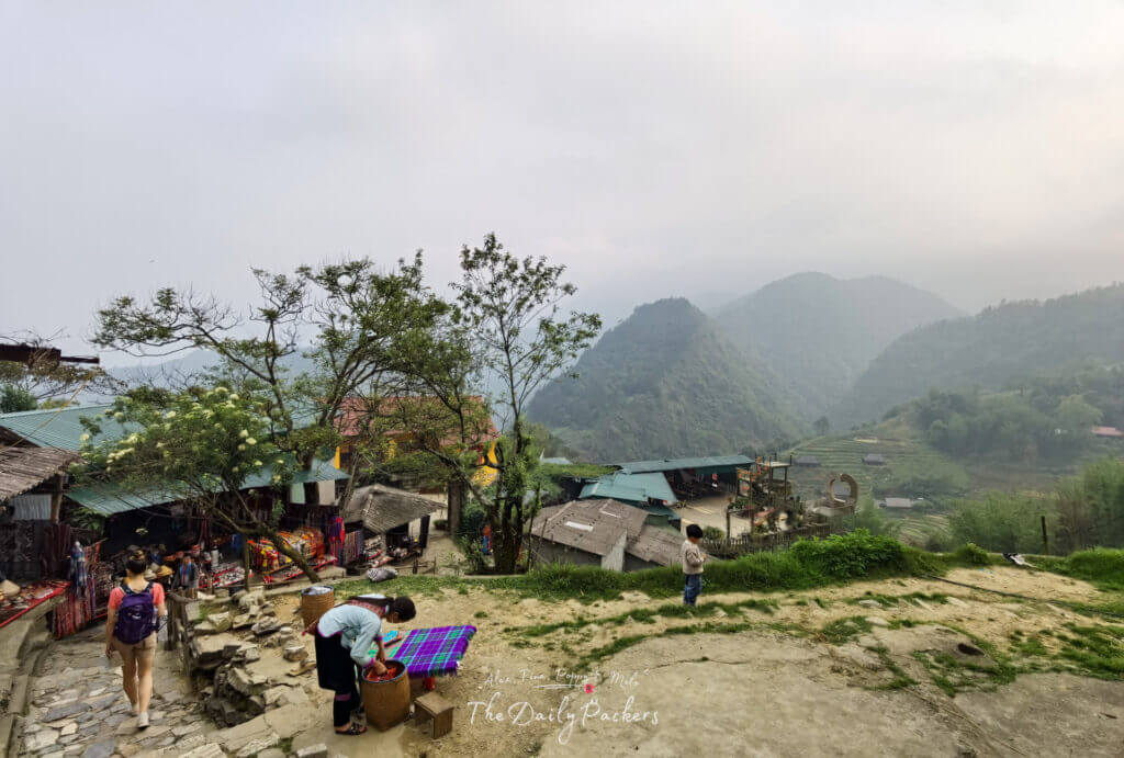 Woman walking through Cat Cat Village as a local woman works on textiles and a child stands nearby, with mountain views in the background.