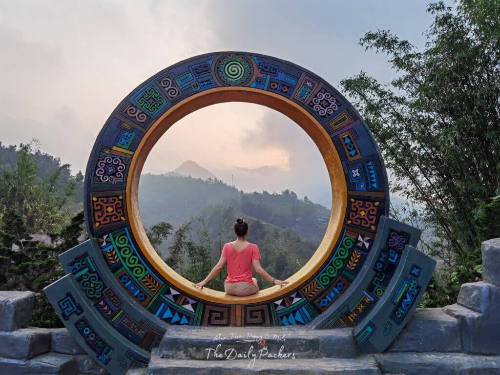 Woman sitting inside a colorful circular viewpoint frame overlooking misty mountains in Cat Cat Village.