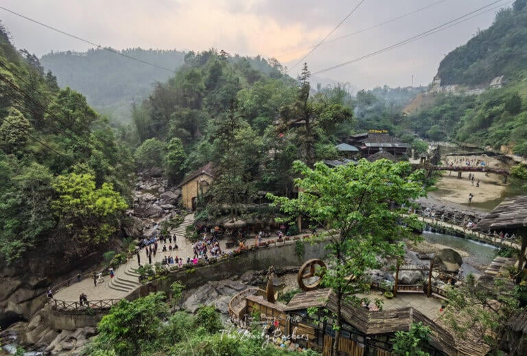 Aerial view of Cat Cat Village’s main square surrounded by forested hills, shops, and a river.