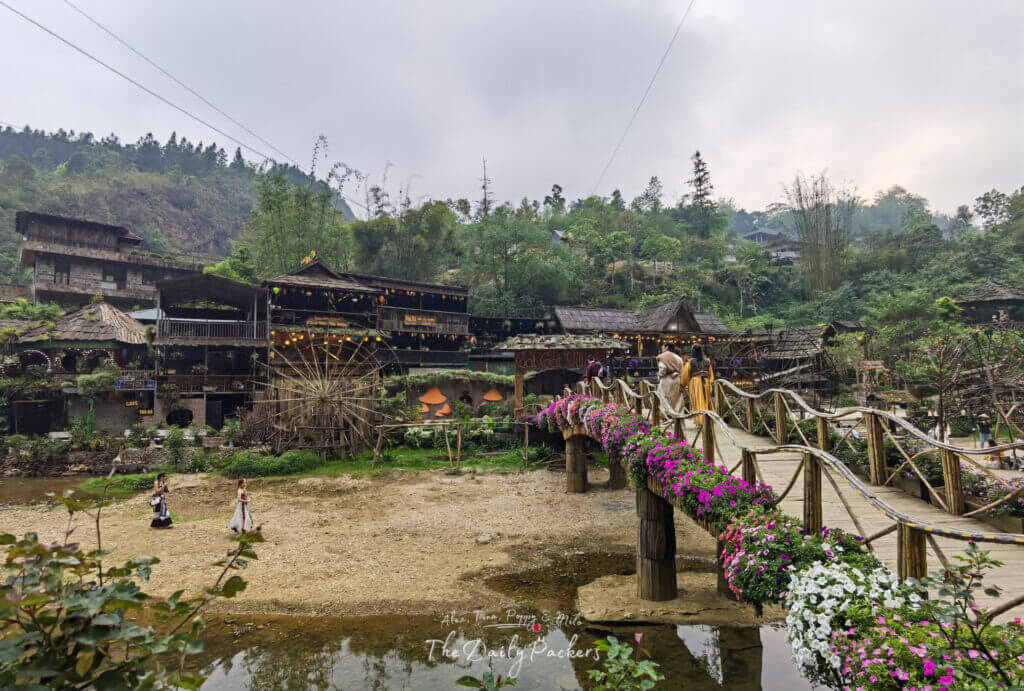 Flower-decorated wooden bridge in Cat Cat Village leading to rustic wooden houses and shops.
