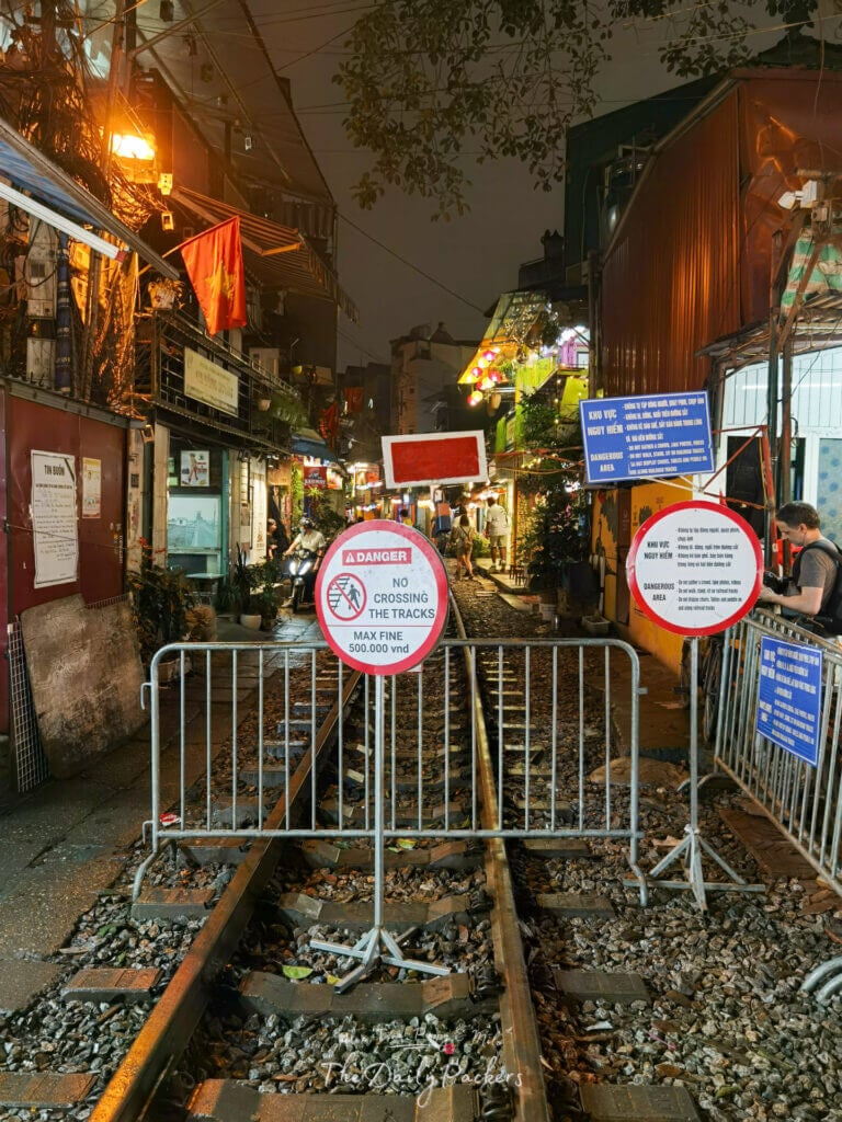 Vue nocturne de l'entrée de la rue du train de Hanoï, bloquée par des barrières et des panneaux d'avertissement, avec des cafés illuminés en arrière-plan.