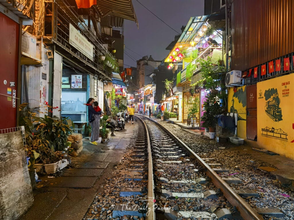 Vue nocturne de la rue du train de Hanoï avec des lumières brillantes, des œuvres d'art murales et un couple debout à côté des voies ferrées.
