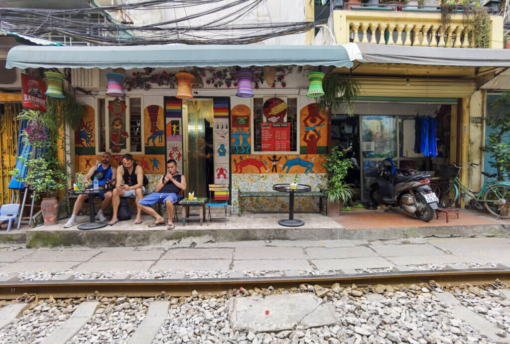 Vue de jour d'un café coloré de la rue du train de Hanoï, avec des peintures murales, des lanternes suspendues et des gens qui se détendent près des rails.