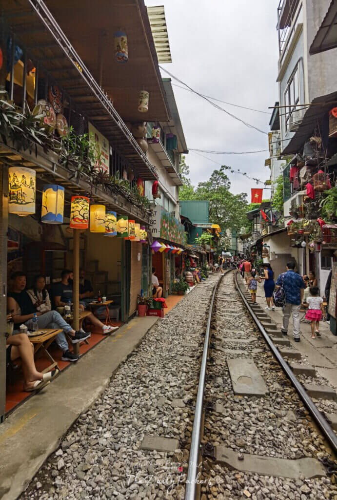 Vue verticale de la rue du train de Hanoi pendant la journée, avec des cafés des deux côtés et des gens qui marchent le long de la voie ferrée.