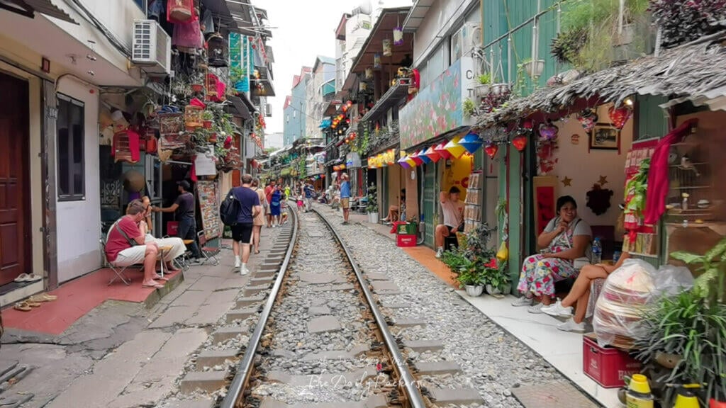 Rue du chemin de fer à Hanoi avec des touristes et des habitants assis à l'extérieur des cafés et des magasins alors qu'un train approche au loin au crépuscule.
