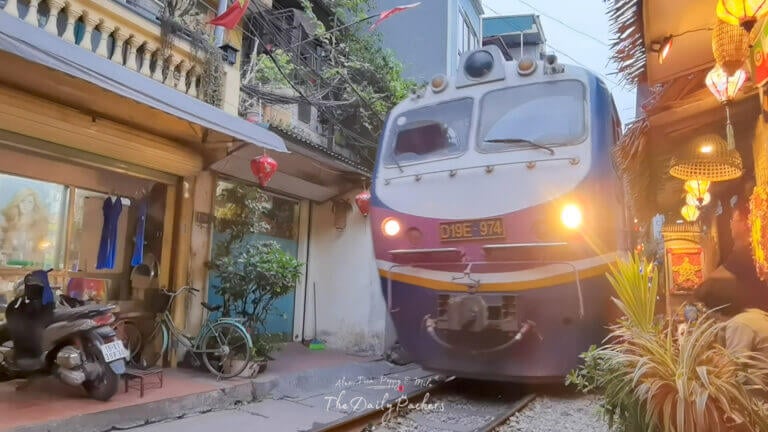 Locomotive approaching closely on Hanoi Train Street, its bright headlights contrasting with the warmly lit shops and decorations nearby