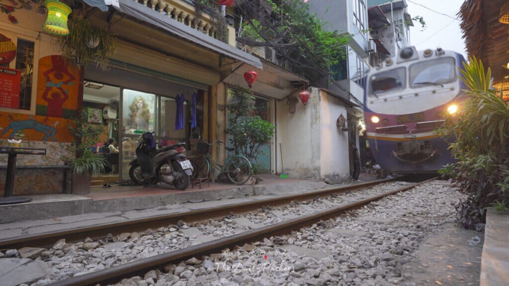 Train traversant la rue des trains de Hanoi le soir, avec des boutiques, des lanternes et des motos le long de l'étroite voie ferrée.