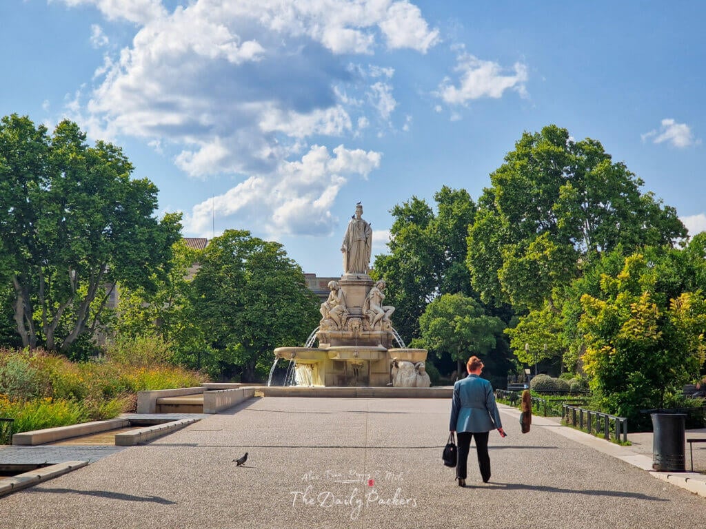 Statue de Pradier surplombant une fontaine, entourée de verdure, lors d'un après-midi ensoleillé à Nîmes.