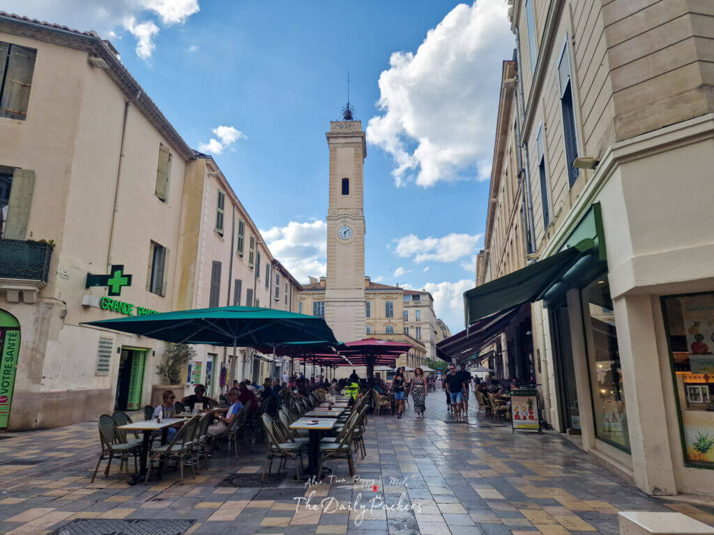 Rue bordée de cafés à Nîmes menant à la tour de l'horloge centrale sous un ciel bleu