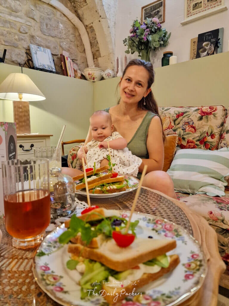 Femme souriante avec son bébé au Cottage Annabelle à Nîmes, assise à une table vintage garnie de sandwichs et de boissons.