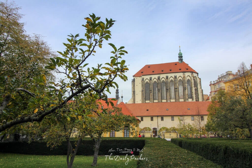 View of the Gothic-style Church of Our Lady of the Snows in Prague, framed by trees and garden greenery.