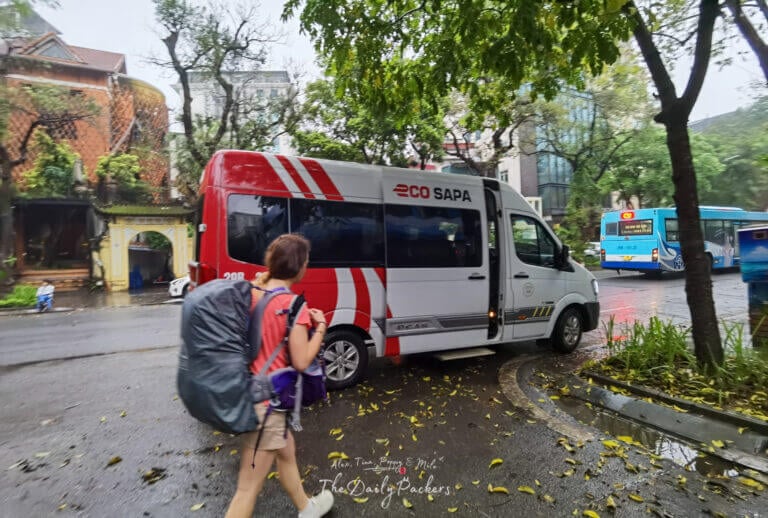 Traveler with a large backpack walking toward an Eco Sapa minivan on a rainy street in Vietnam.