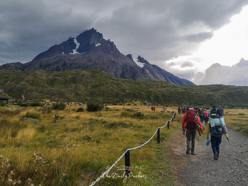 Hikers with backpacks walking from Paine Grande towards Refugio Grey under dramatic mountain clouds.