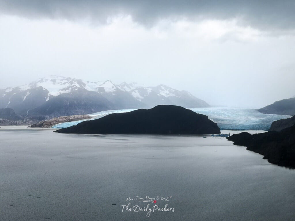Vue du Lago Grey avec le glacier Grey au loin, entouré de montagnes escarpées sous un ciel nuageux de Patagonie.