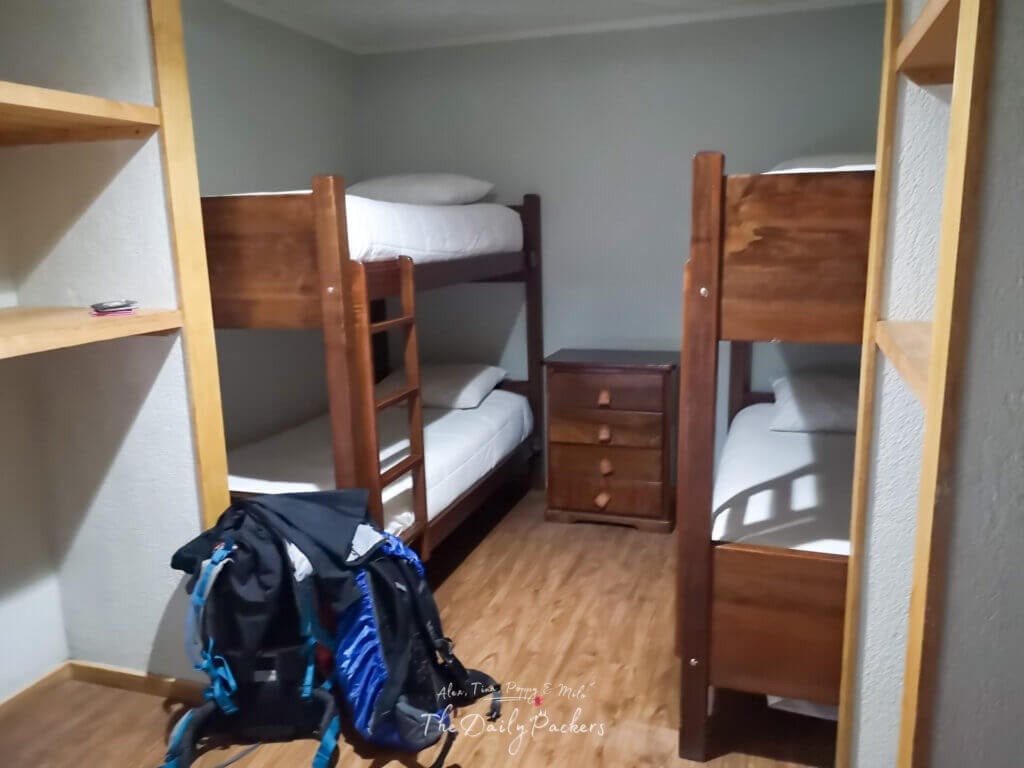 Refugio Grey dorm room with two bunk beds, wooden shelves, and backpacks on the floor.
