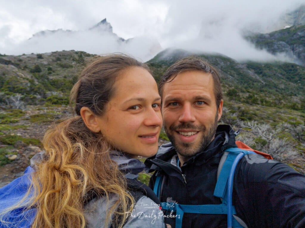Close-up selfie of a smiling couple in hiking gear with mist-covered mountain peaks in the background.