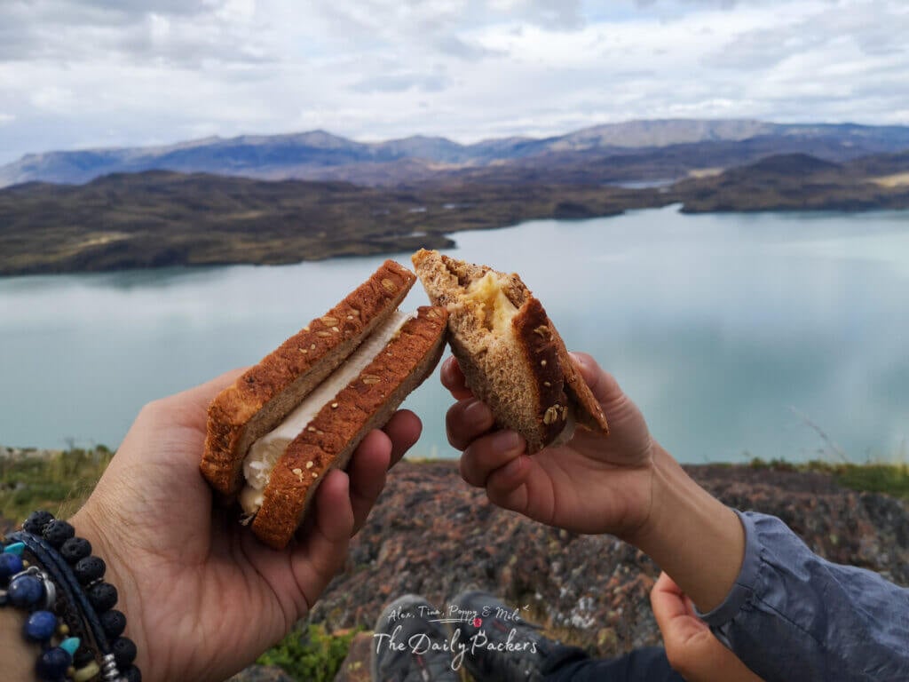 Two hands holding sandwiches with a turquoise lake and rugged Patagonian landscape in the background.