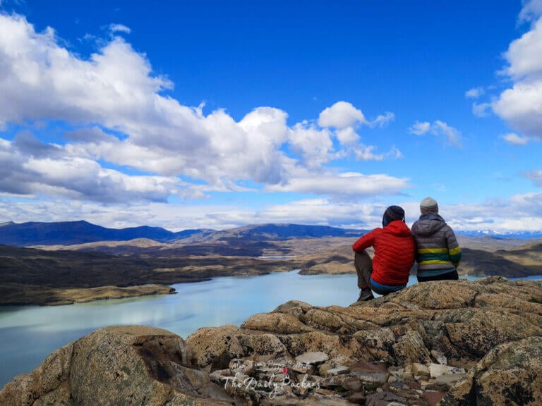 Couple in hiking gear sitting together on a rocky summit, smiling at the camera with sweeping views of Lake Nordenskjöld during the W-Trek day 4