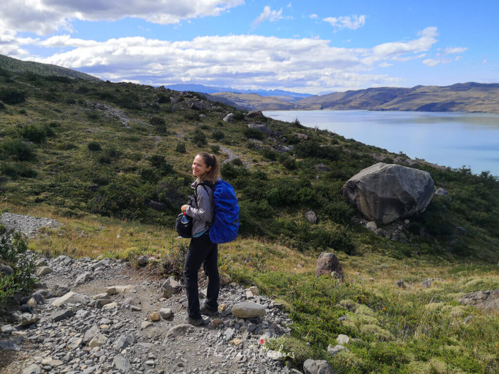 Female hiker with a large blue backpack smiling on a mountain trail overlooking the turquoise waters of Lake Nordenskjöld.