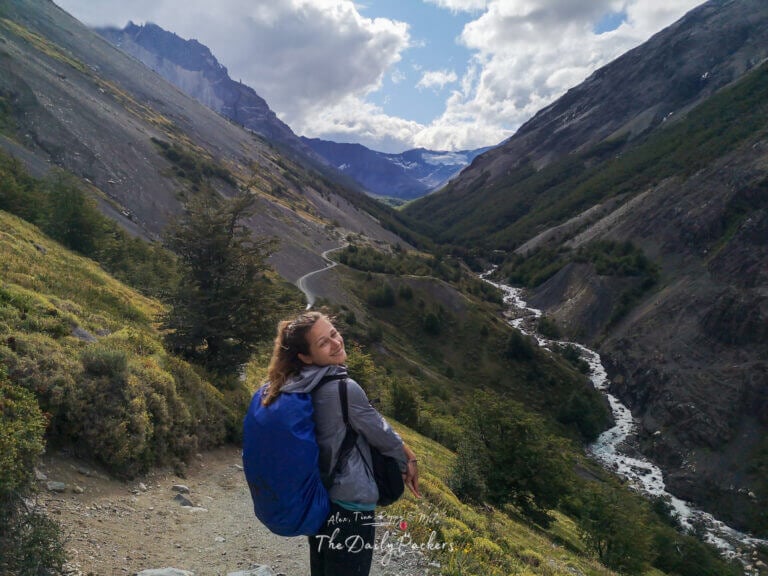 Hiker smiling while sitting on a mountain trail overlooking a winding river and steep valley slopes in Torres del Paine National Park.
