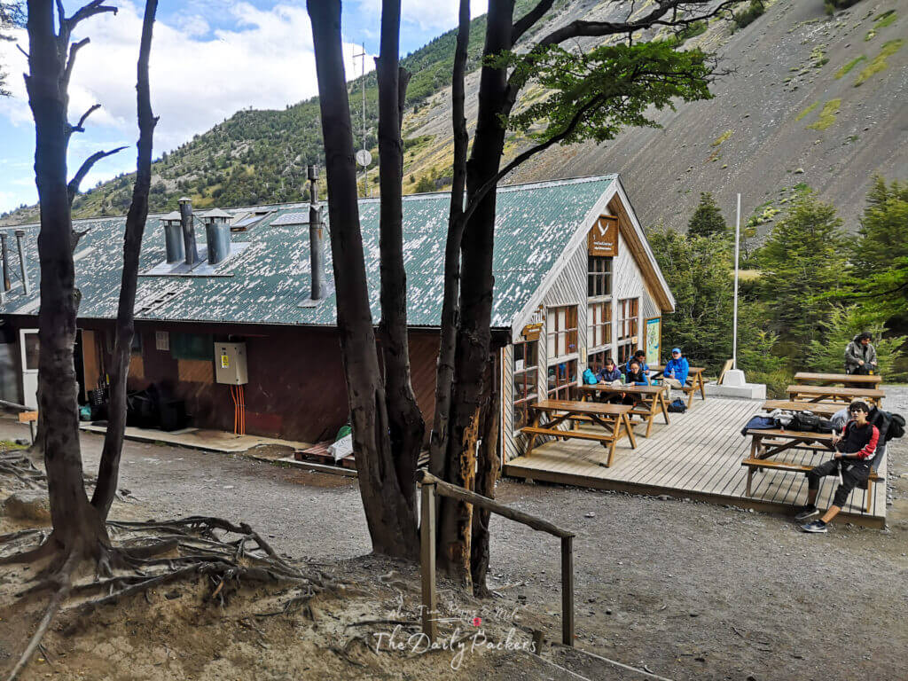 Refugio Chileno shelter building with hikers sitting at wooden picnic tables on a deck surrounded by mountain scenery.