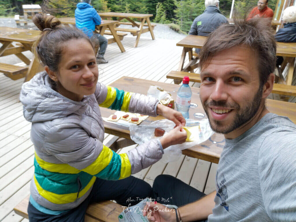 Couple enjoying snacks at an outdoor wooden picnic table at Refugio Chileno, surrounded by other hikers.