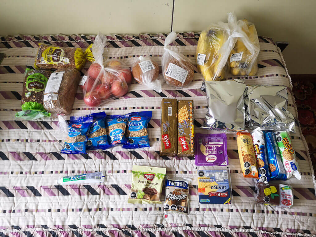 Food supplies laid out on a bed for trekking—bread, fruit (apples and bananas), nuts, crackers, cookies, cheese, sweets, and other snacks.