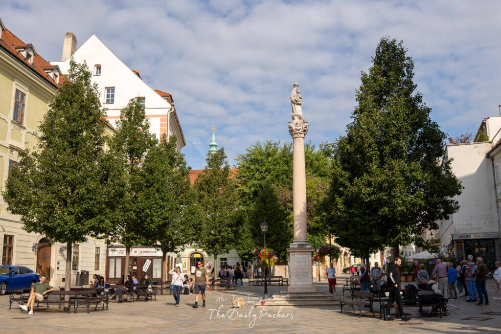 Marian Column statue in Bratislava’s old town square surrounded by trees and people.