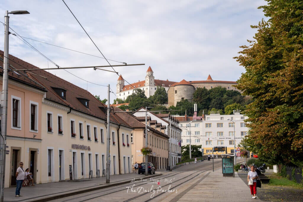Street view leading up to Bratislava Castle, showing tram lines, pastel-colored houses, and the castle in the background.