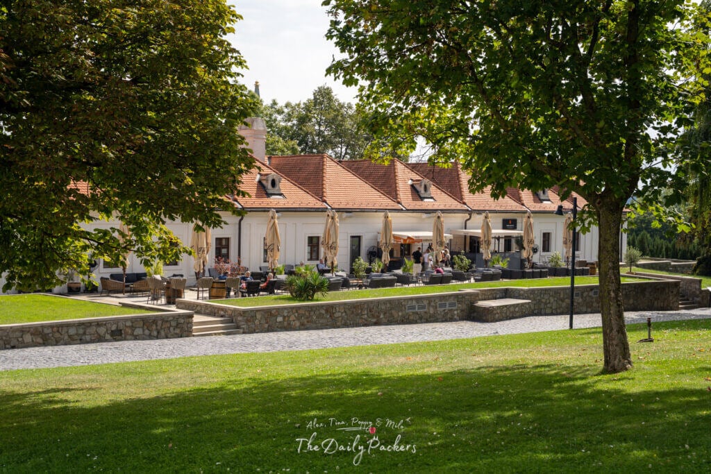 Outdoor restaurant terrace next to Bratislava Castle, with people dining under umbrellas in a green park setting.
