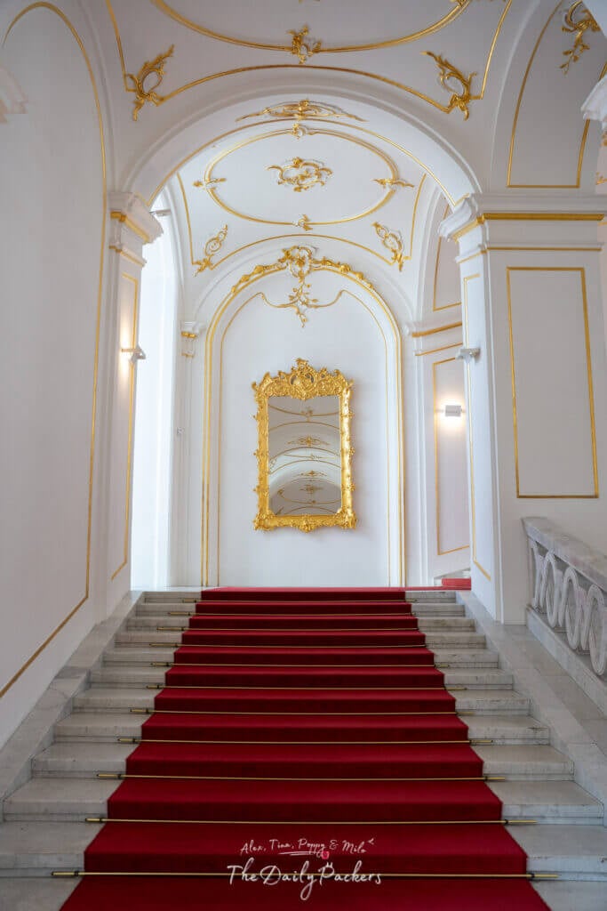 Grand marble staircase with a red carpet and a golden framed mirror inside Bratislava Castle.