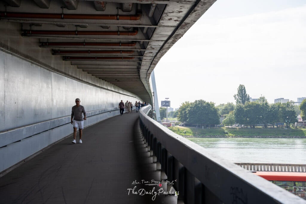 Pedestrian walkway under the SNP Bridge leading to Bratislava UFO Tower, with people walking along the Danube.
