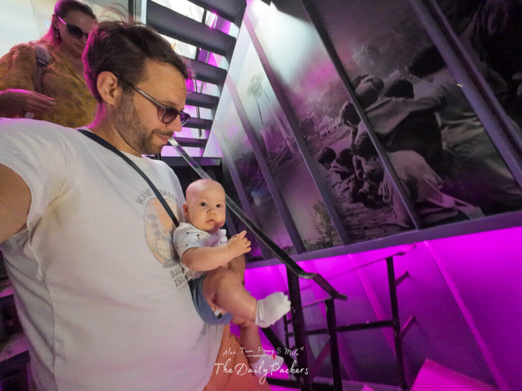 Father carrying baby while descending the purple-lit staircase inside the Bratislava UFO Tower, with Tina following behind.