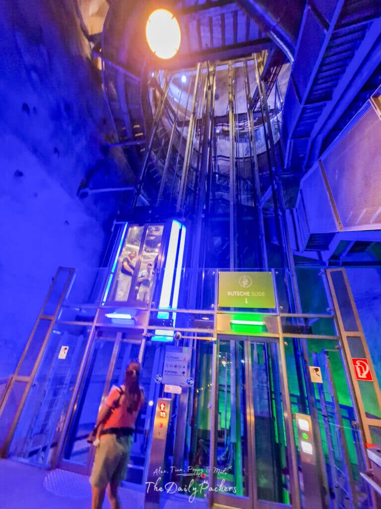 À l'intérieur du tunnel illuminé du Schlossberglift à Graz, avec des lumières colorées se reflétant sur l'ascenseur en verre et l'entrée du toboggan.