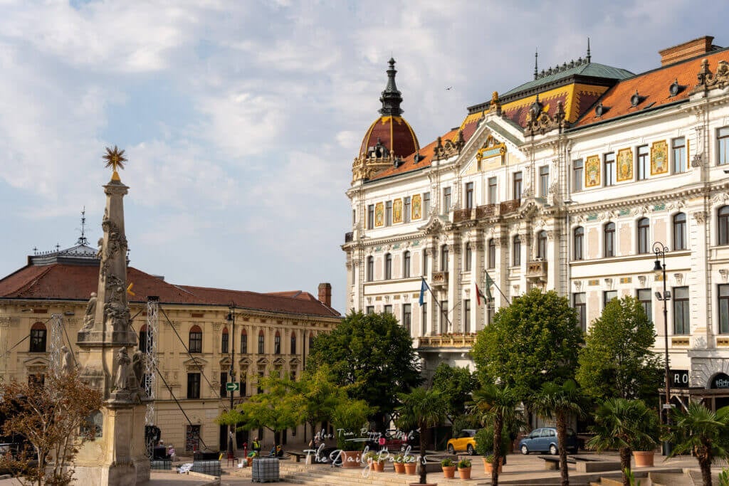 Széchenyi Square in Pécs with the Holy Trinity statue and grand baroque buildings under a partly cloudy sky.