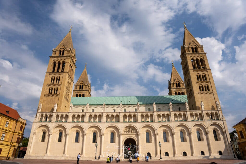 Front view of Pécs Cathedral with its four tall towers and green roof against a partly cloudy sky.