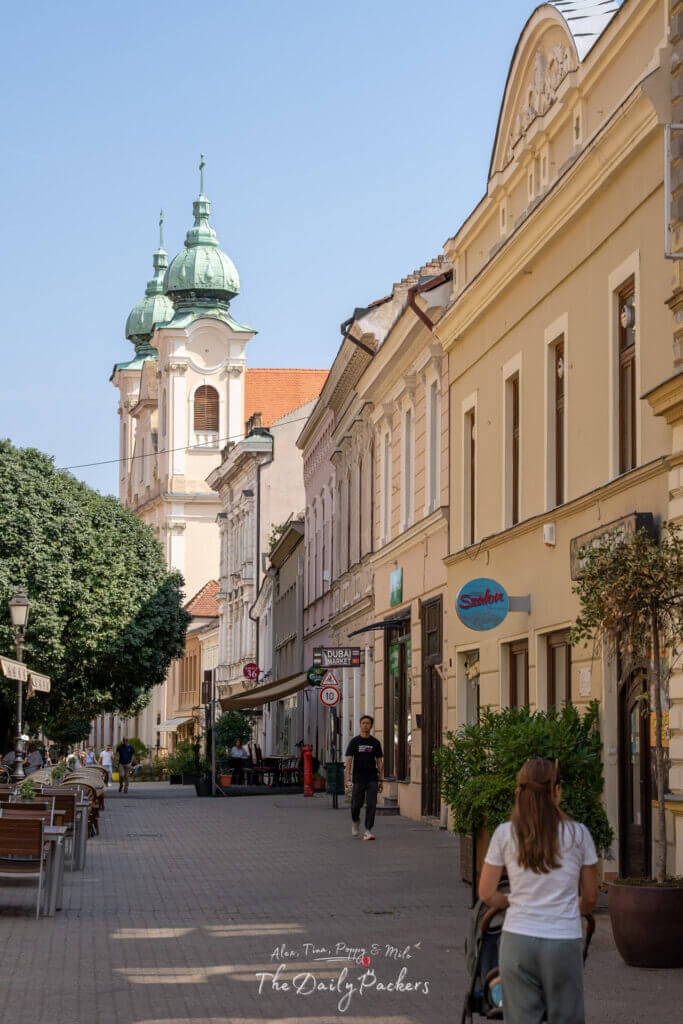 View of Pécs’ bustling main street with shops, cafes, and the twin green-domed church tower in the background.