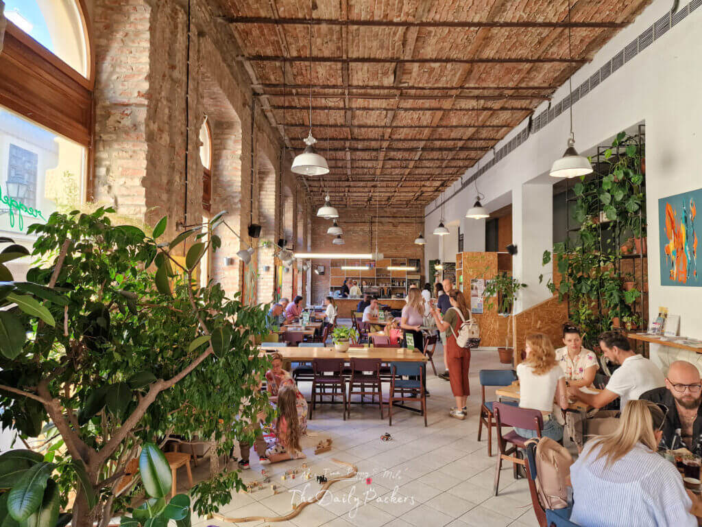 Interior of Reggeli café in Pécs, featuring exposed brick walls, plants, and people enjoying food and drinks in a lively atmosphere