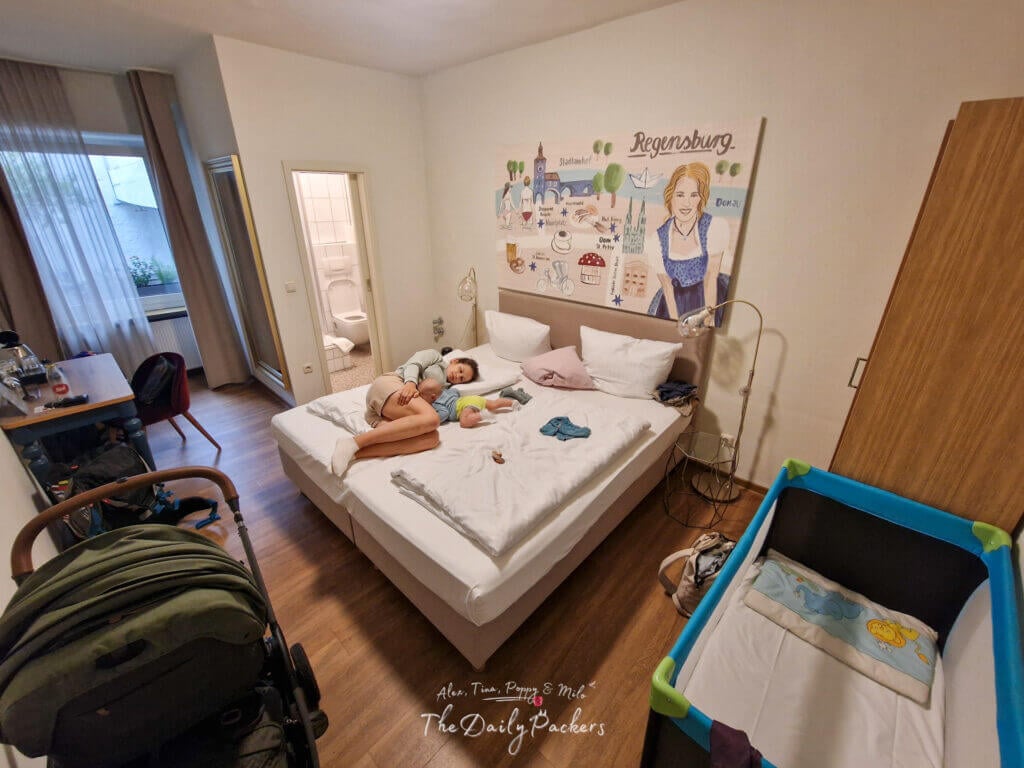 Hotel room in Regensburg with a family resting on the bed, crib, stroller, and Regensburg-themed wall art.